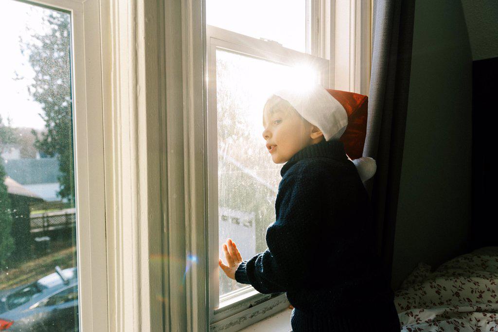 Little boy with Christmas hat looking out the window for Santa clause