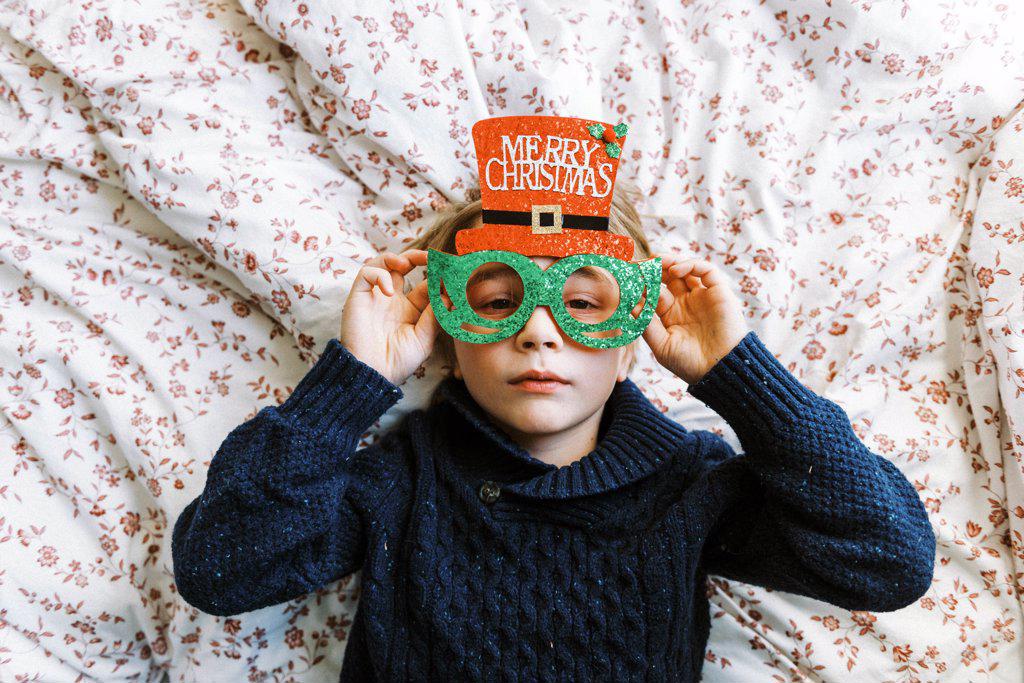 Little boy in a knit sweater with Christmas glasses lying on bed