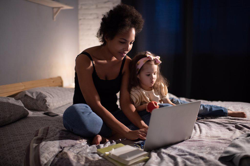 Ethnic mother using laptop for work near playing daughter