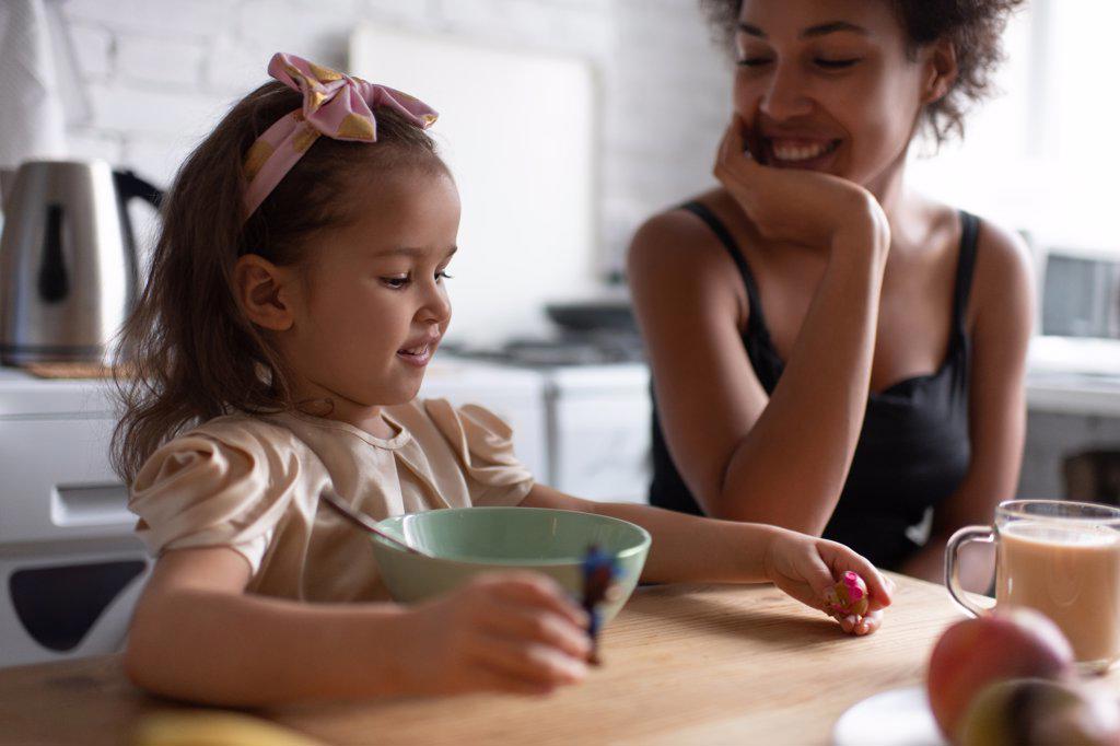Cheerful ethnic girl playing during breakfast with mother