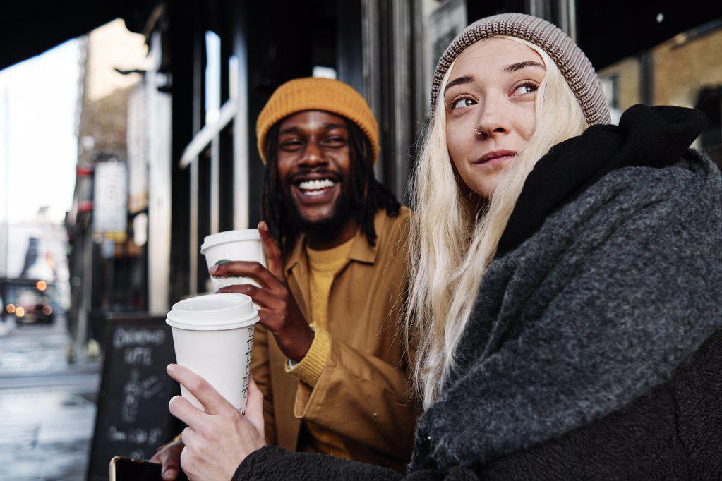 multiracial couple sitting outdoors laughing while drinking coffee. biracial lovers on a date concept.