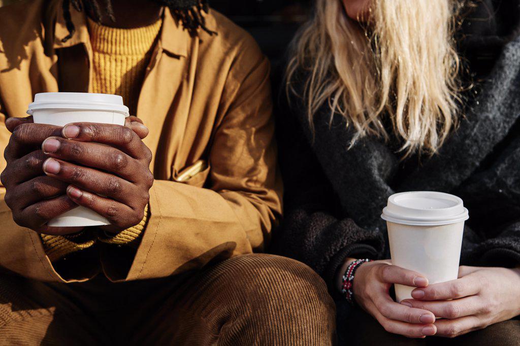 multicultural unrecognizable friends holding a paper cup of coffee while sitting together. focus on the hands.