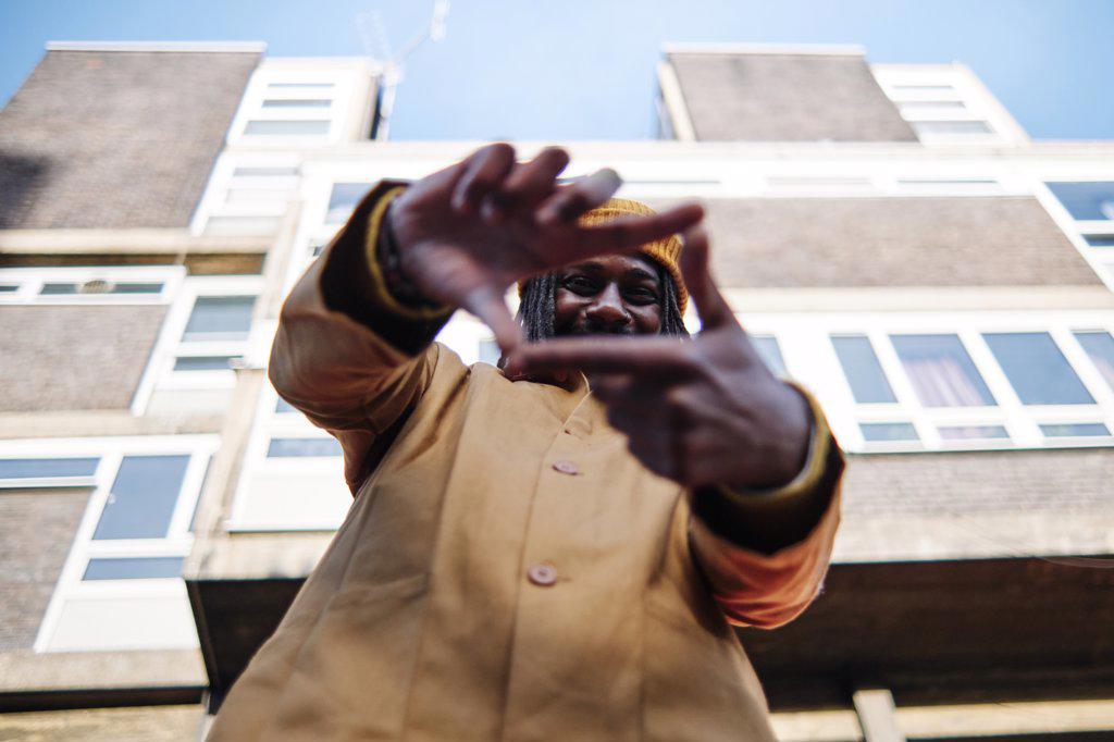 young african guy looking at camera, smiling through finger frame while standing in front of a building