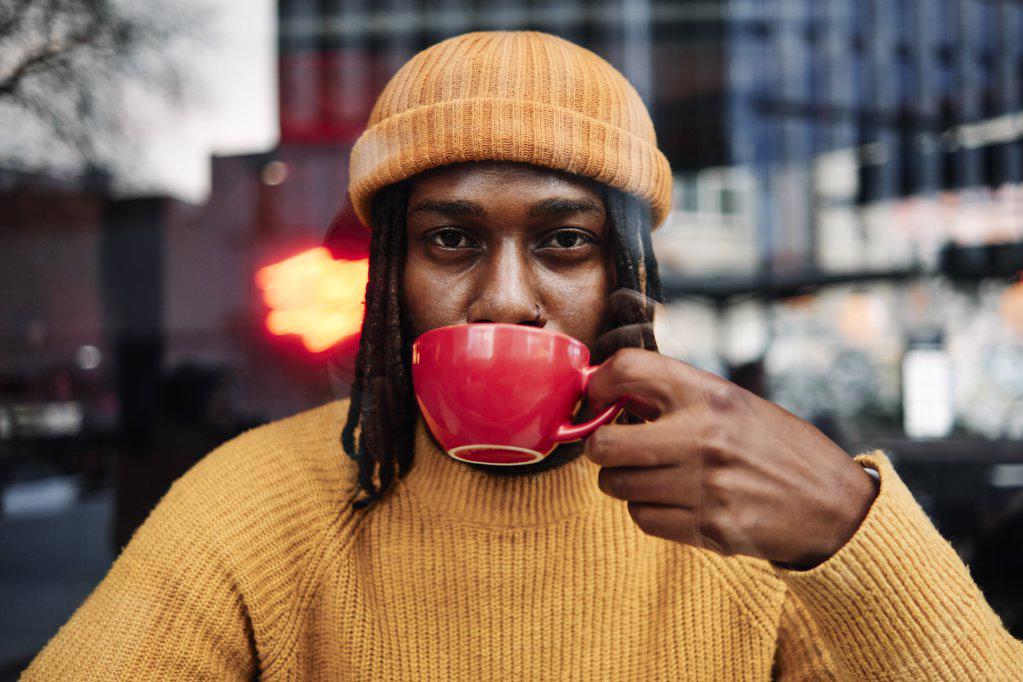 young african guy staring at camera as he drinks a cup of coffee at a cafe, reflection of the city on window