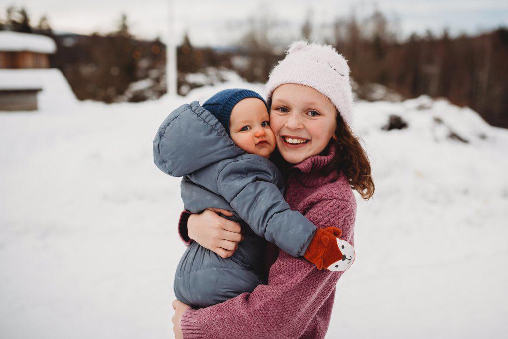 Smiley Big sister holding baby brother outside in the snow on cold da
