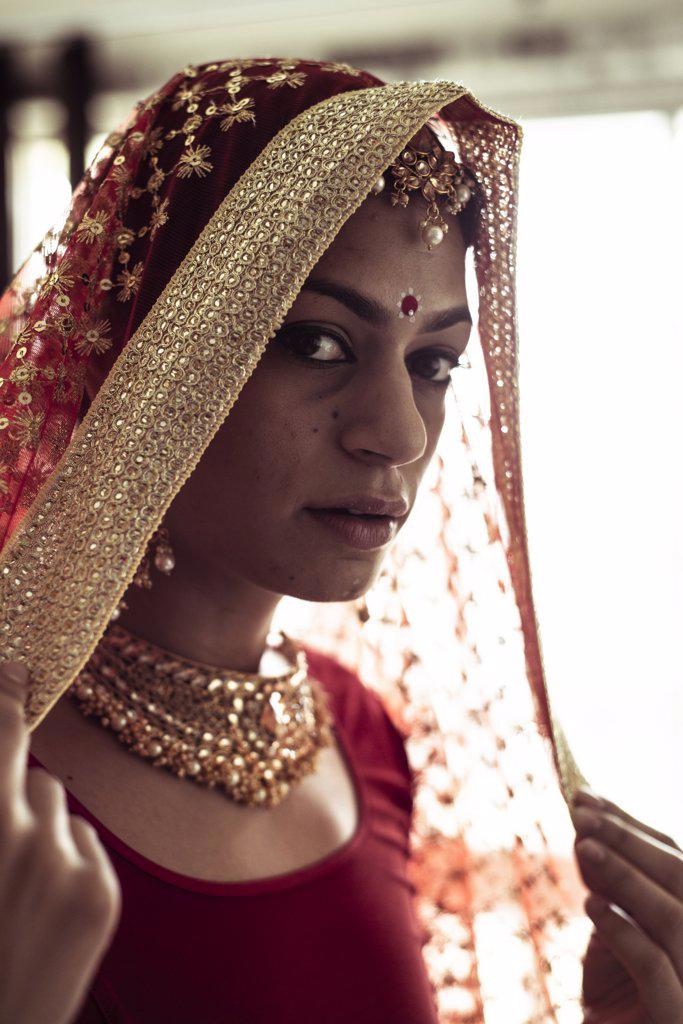 Young Indian woman pulls on red wedding veil in window light