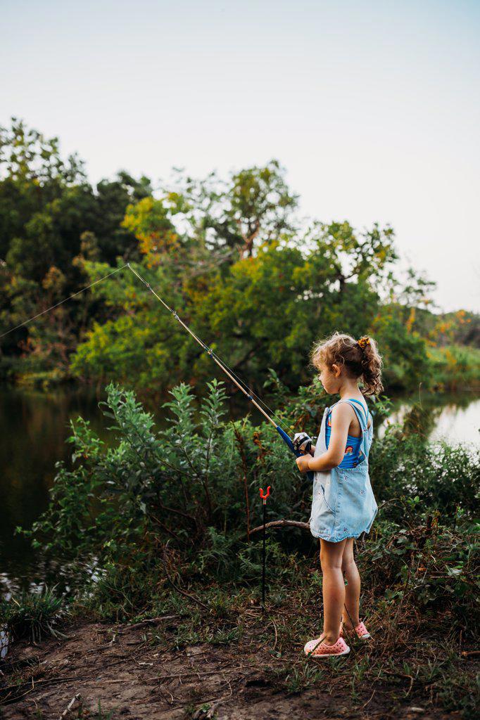 Young girl standing at edge of creek holding fishing pole in summer