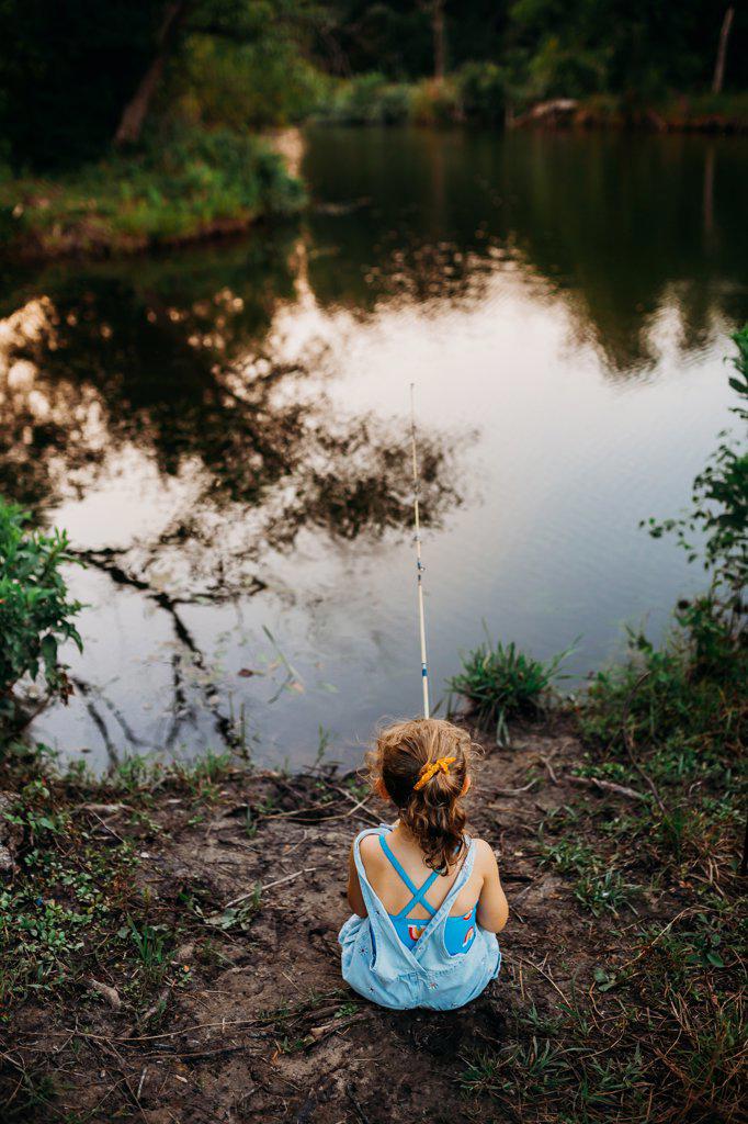 young girl sitting at creek bank fishing at sunset in summer