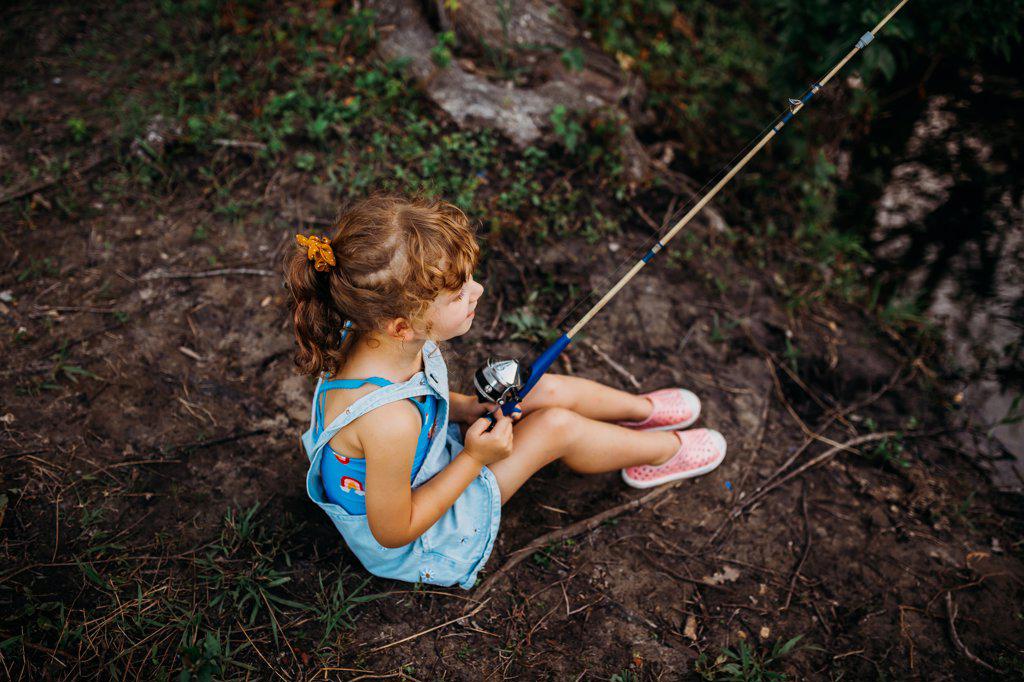 Young girl sitting on ground fishing from creek bank during summer