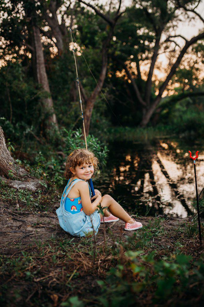 Young girl sitting at creek bank holding fishing pole and smiling