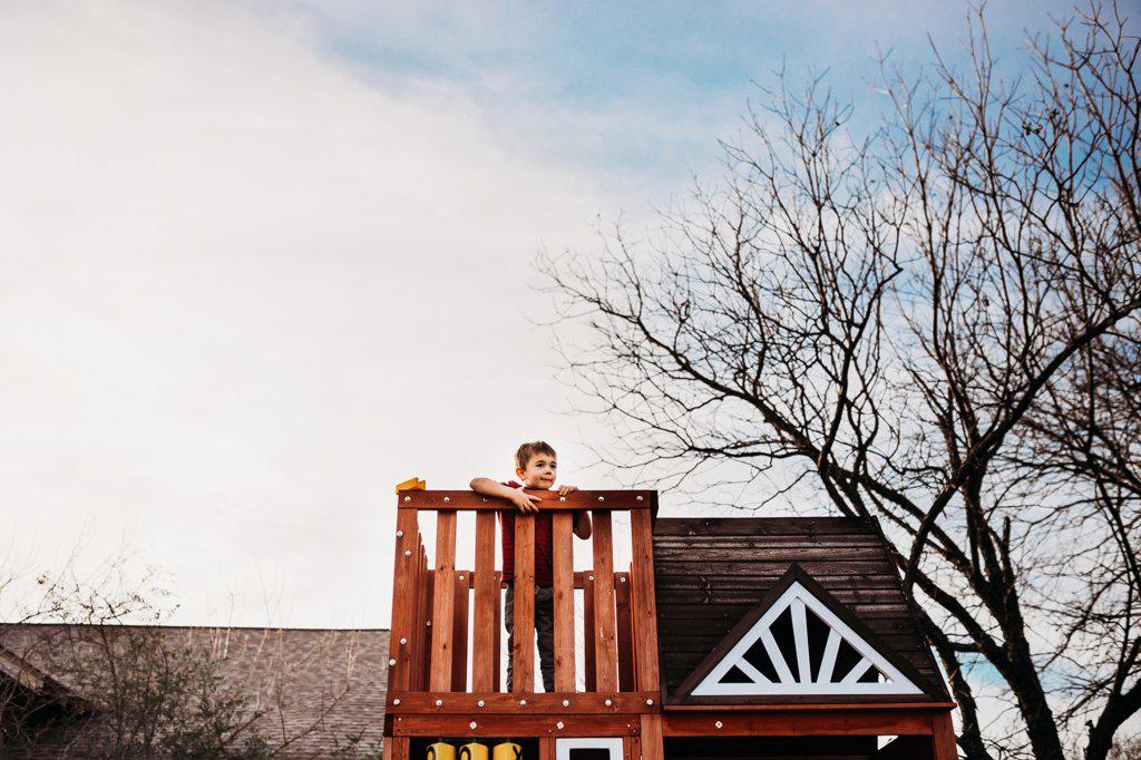 Young boy standing alone in backyard playhouse looking up at sky