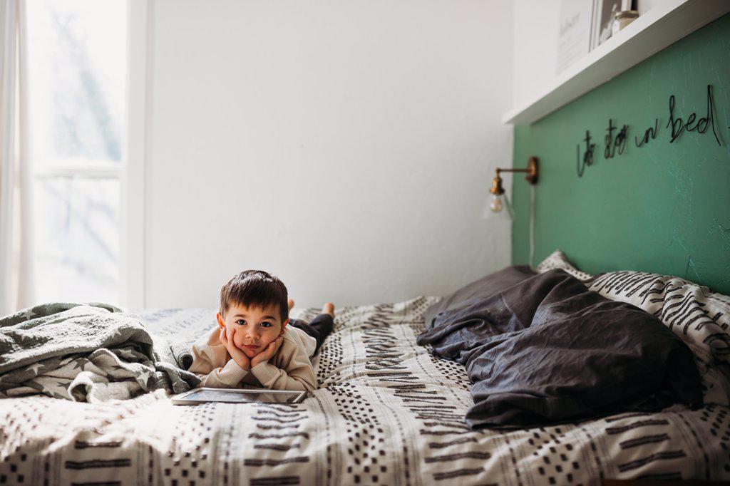 Young boy looking at camera laying on bed with tablet in winter