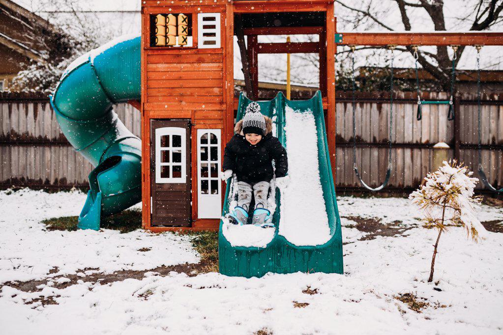 Young boy sliding on snow covered swingset in backyard at home