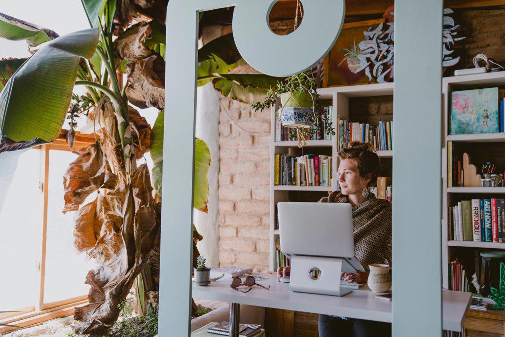 Woman works on laptop from home office surrounded by books and plants