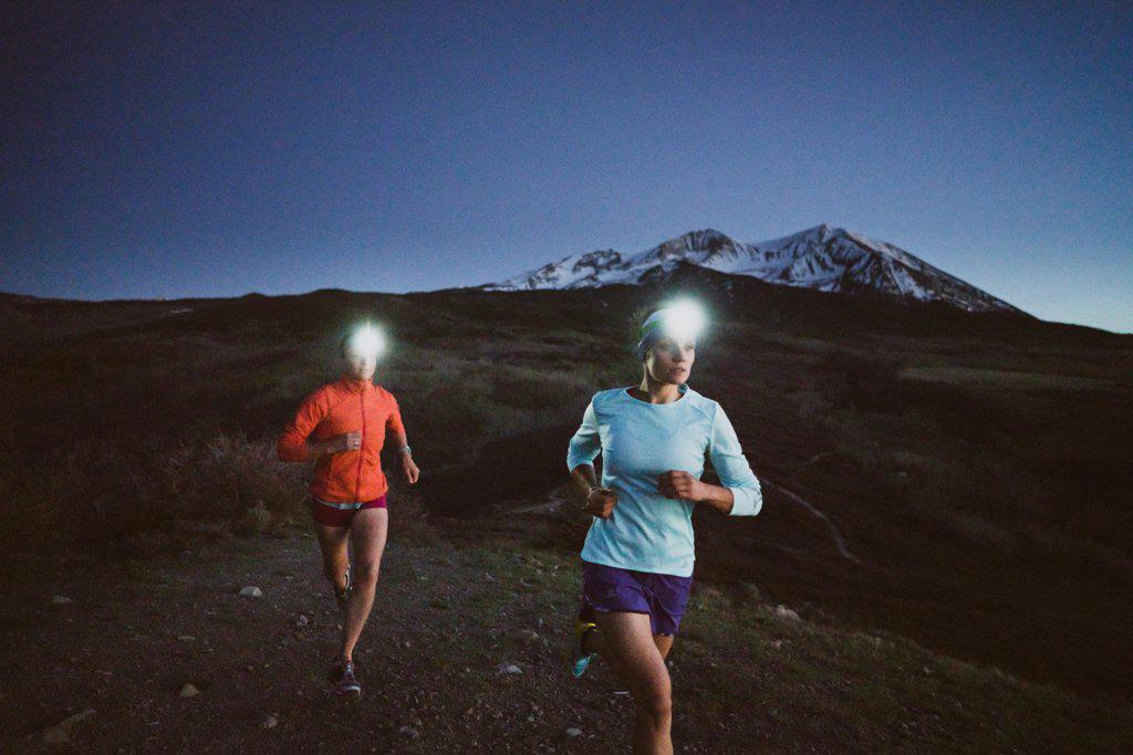 Two women run at night in the mountains wearing headlamps