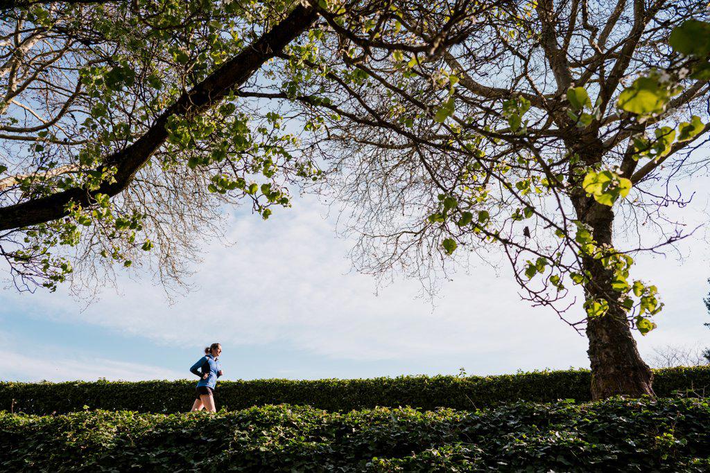 Young woman runs underneath tall green trees and blue skies