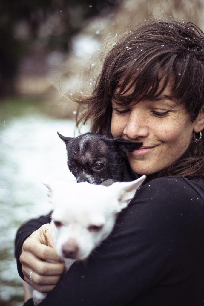 woman with freckles and snow in hair smiles and cuddles two chihuahuas