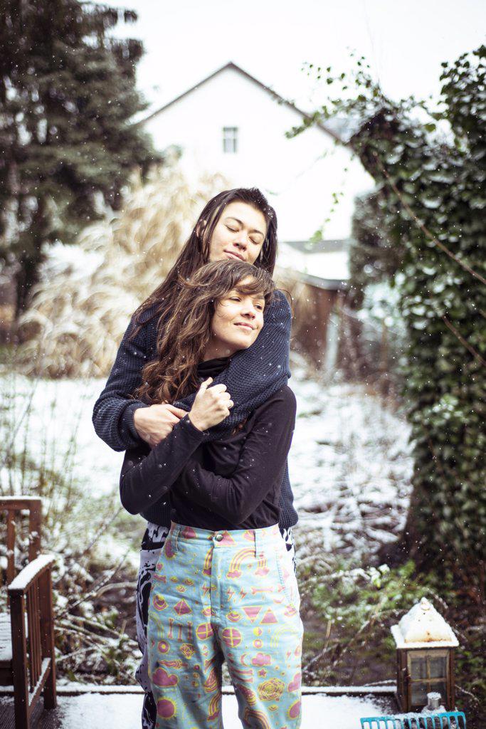 Queer female couple embrace and smile while snow falls in back yard