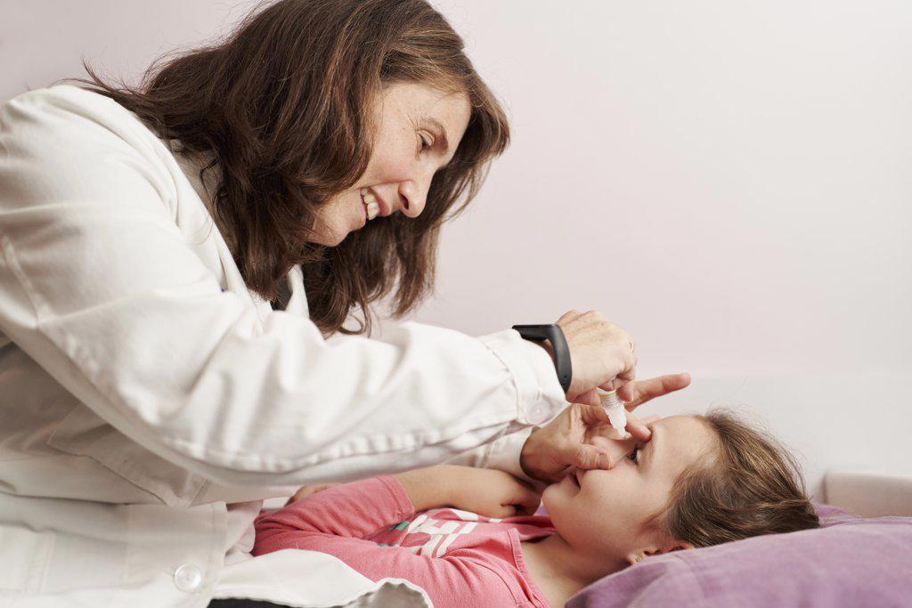 Female doctor smiling and giving eye drops to a little girl in her bed. Home doctor concept