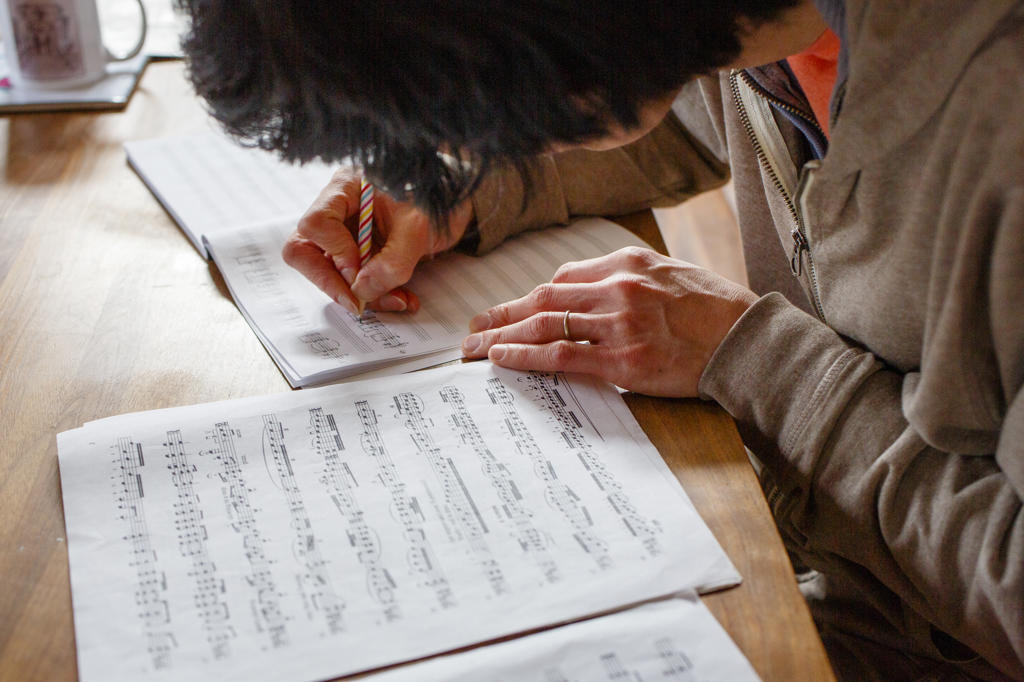 A man sits at a wood table transcribing sheet music with a pencil