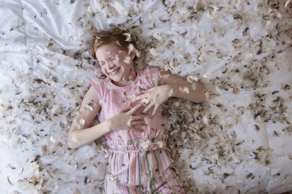 Young red haired girl laying on the bed during a feather pillow fight.