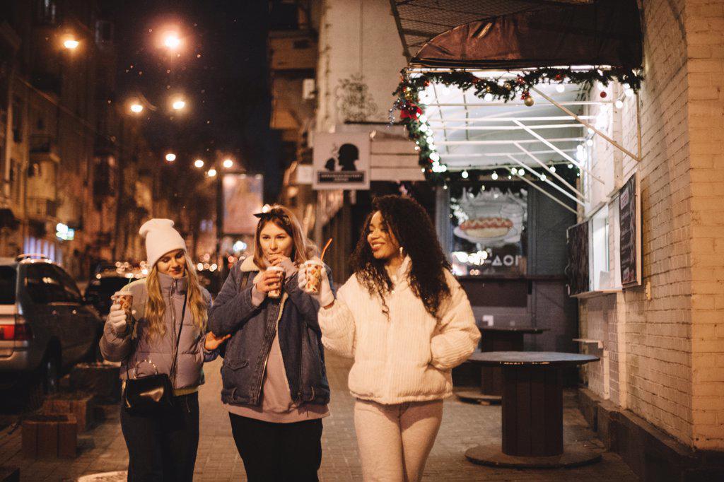 Female friends having hot drinks walking on street in city at night