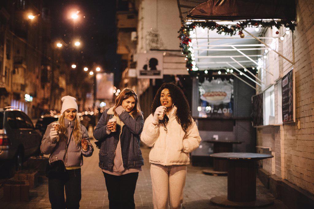 Young women with disposable cups walking on street in city at night
