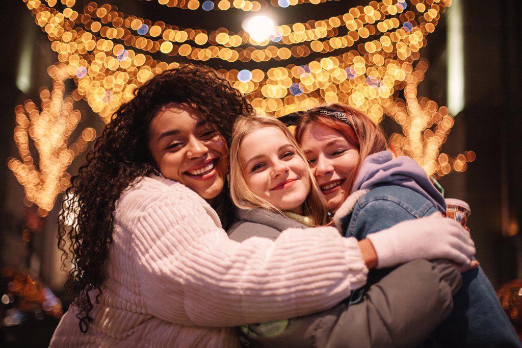 Happy female friends embracing standing by Christmas lights in city