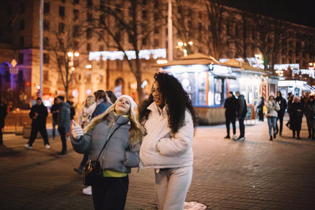 Happy female friends walking on street in city at night during winter