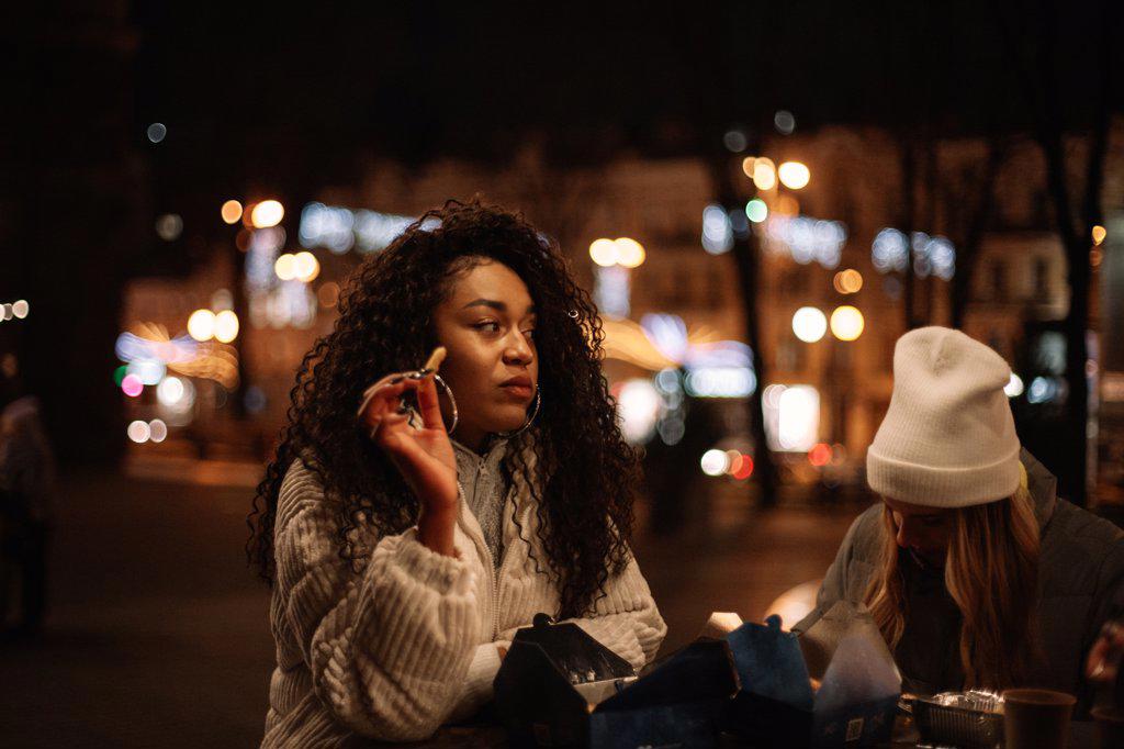 Young women eating street food standing by table in city at night