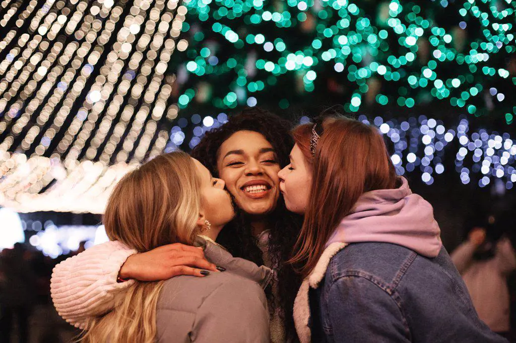 Female friends kissing young woman on the cheeks by Christmas lights