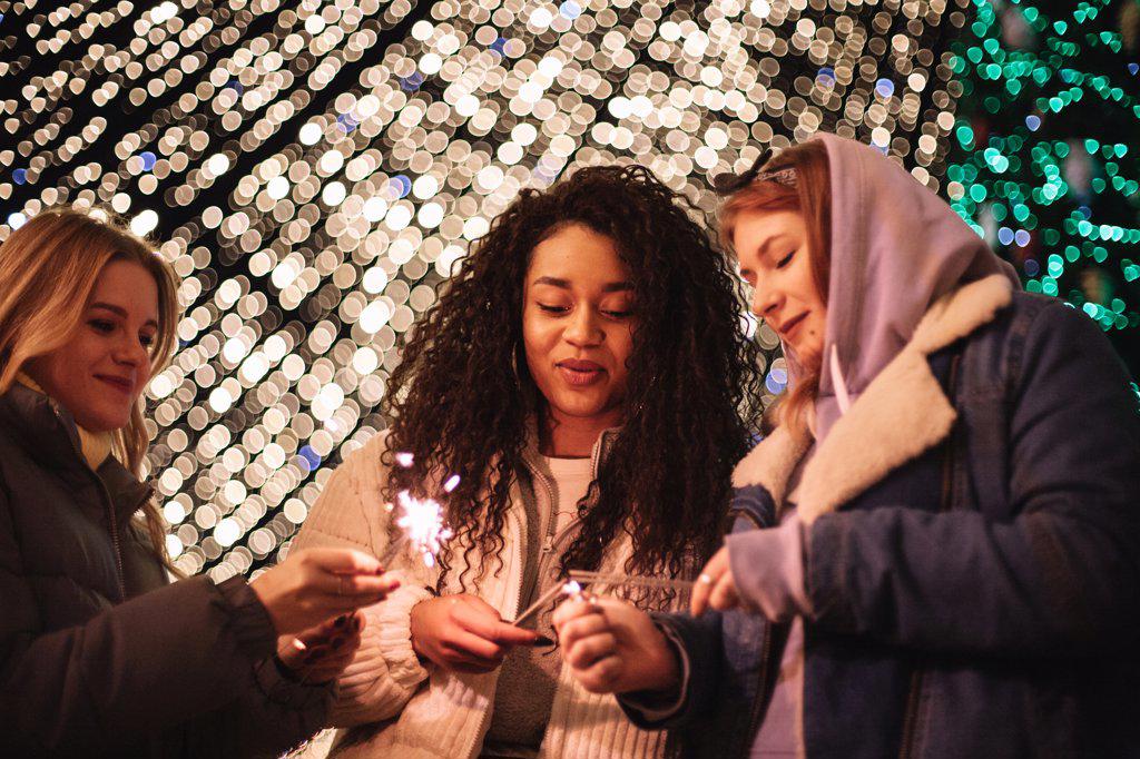 Happy female friends igniting sparklers standing by Christmas lights