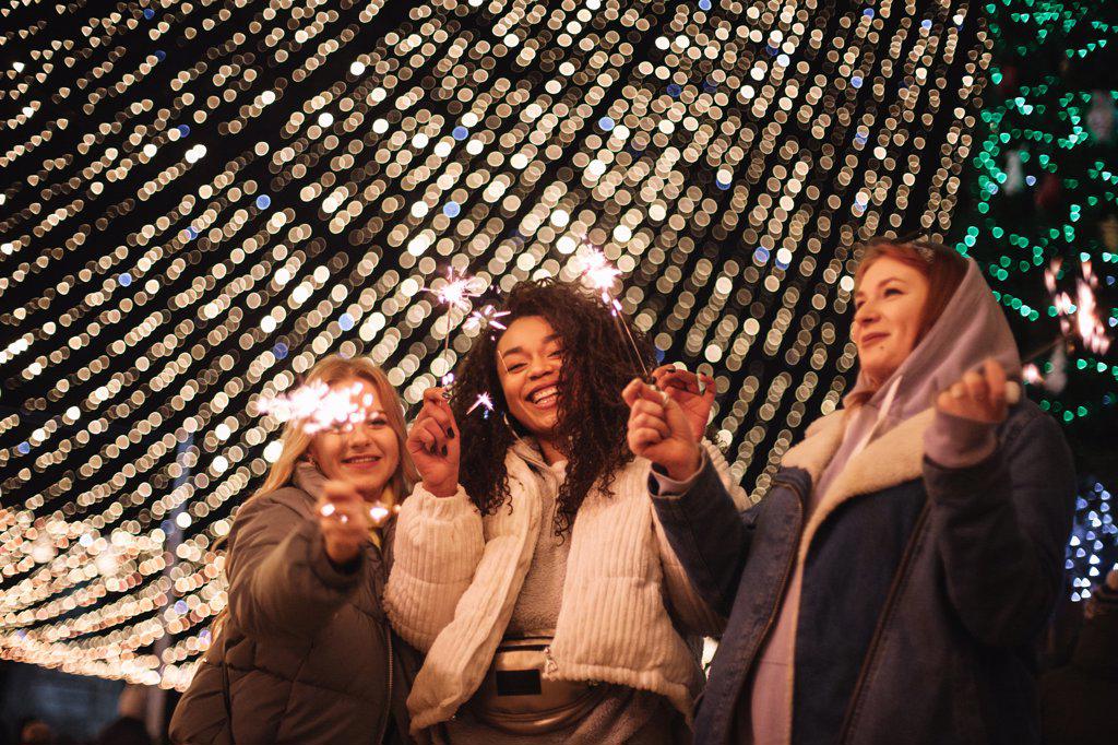 Happy female friends holding sparklers standing by Christmas lights