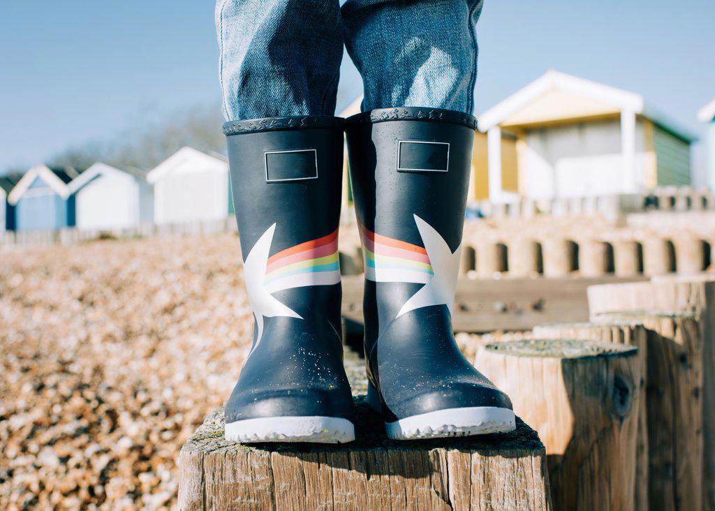 child stood on a wooden post in rain boots at the beach by beach huts