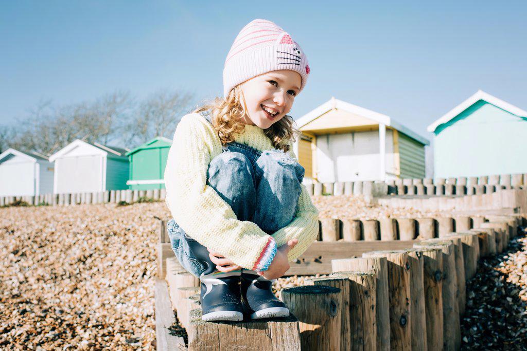 young girl sitting on a pebble beach by colourful beach huts smiling