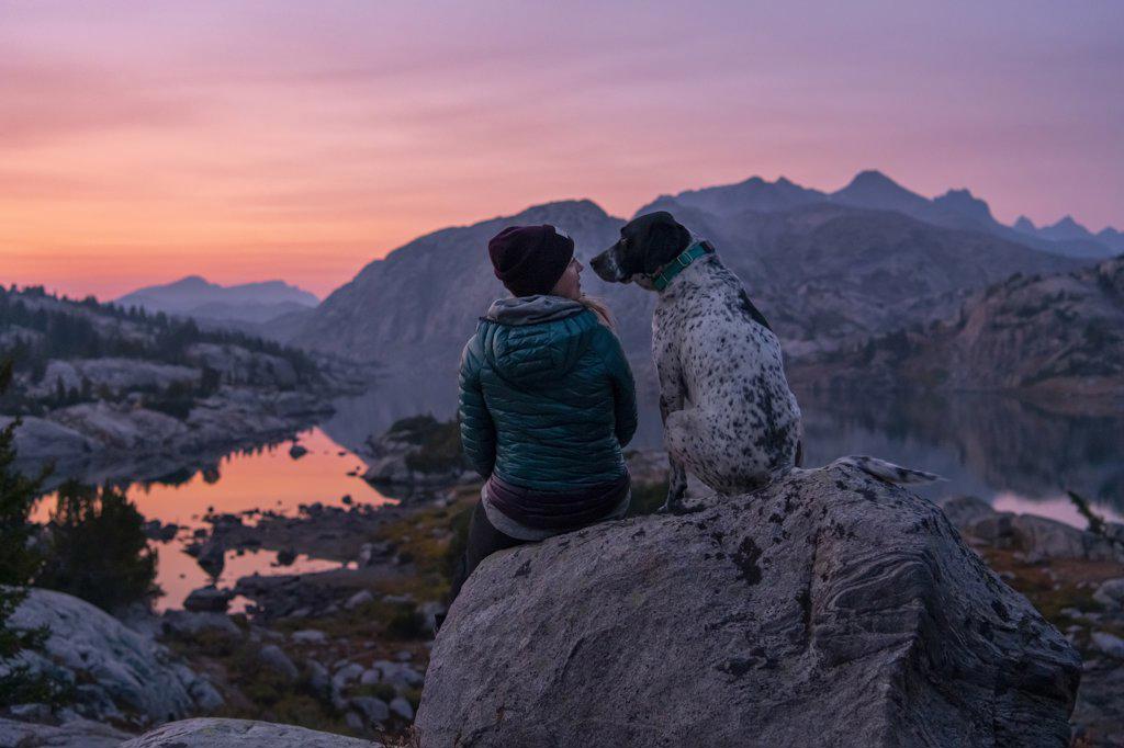 Rear view of young woman sitting on rock with dog on mountain during sunset