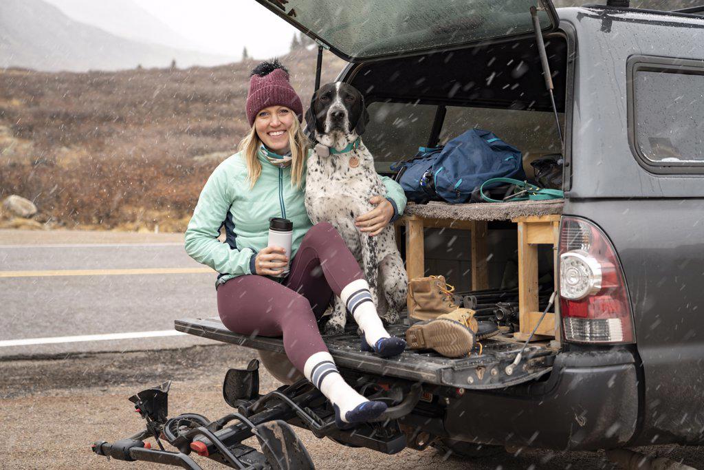 Smiling young woman sitting with dog in trunk of off-road vehicle during snowfall