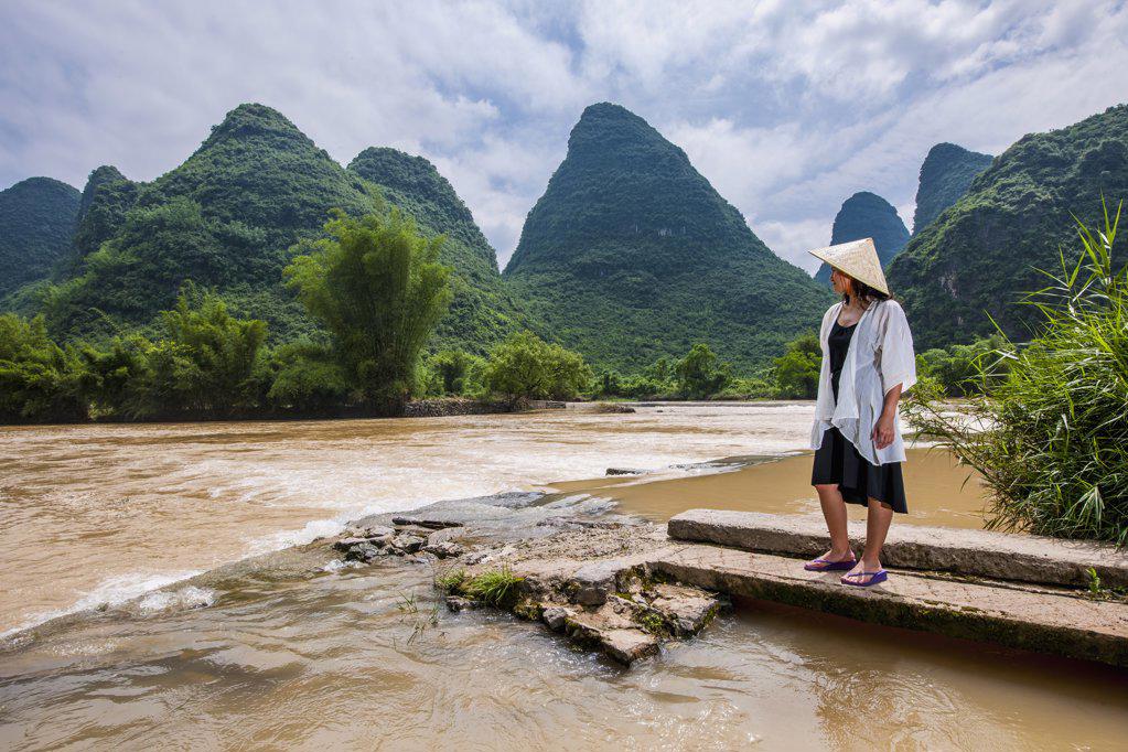 woman in traditional Asian hat standing at river bank in Yangshuo