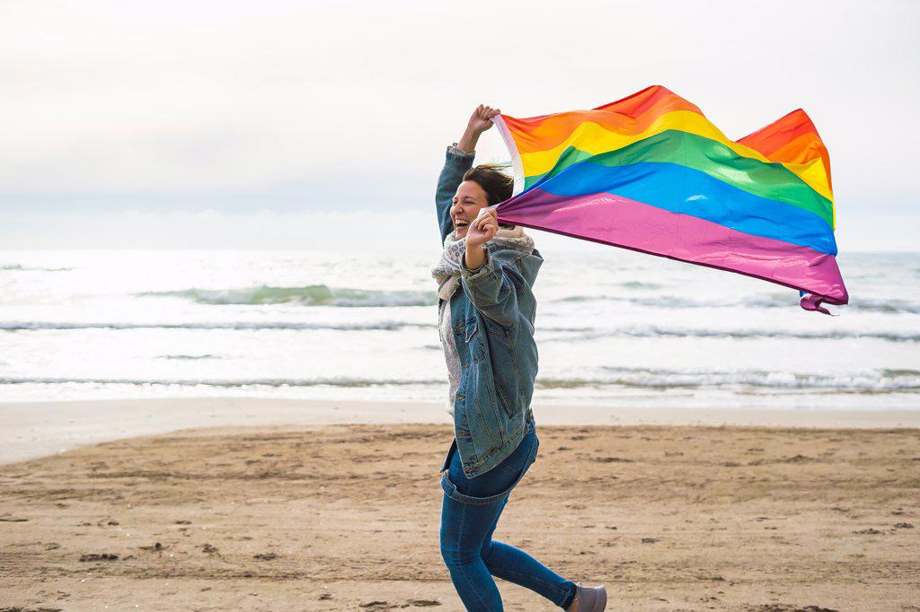 Woman smiling and running with colorful LGBT flag waving on the beach