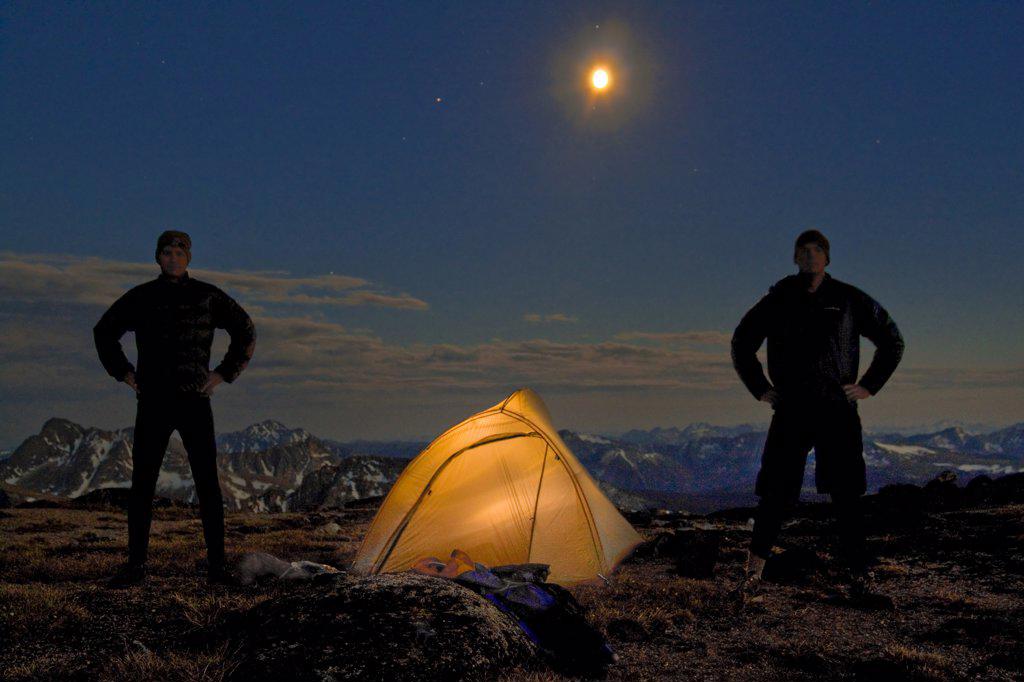 Two men stand next to their lit up tent on a mountain summit.