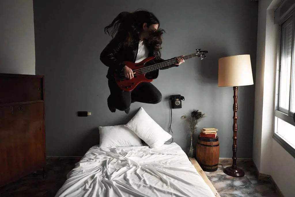 A young man with long hair jumping bed while playing the bass guitar