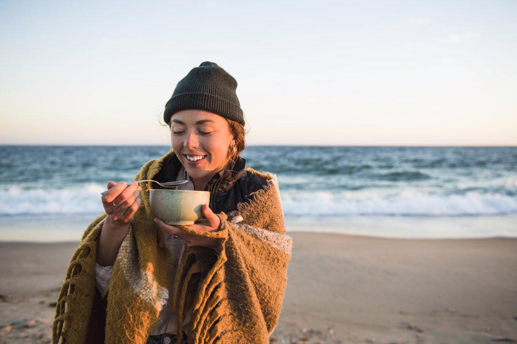 Young woman enjoying bowl of soup while beach car camping in fall