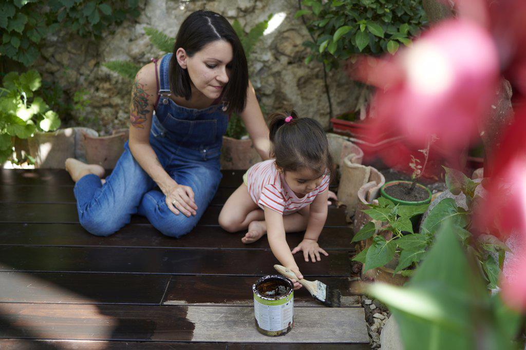 A mother watches as her little girl paints the wood in the garden.