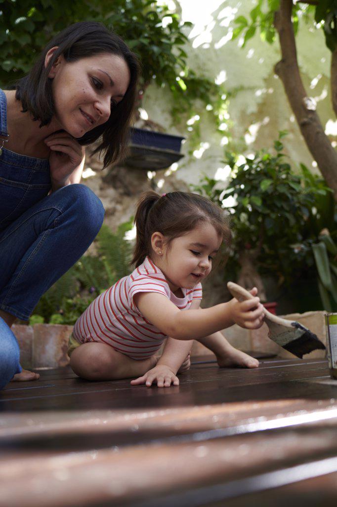 A little girl paints the wood on the floor while her mother watches