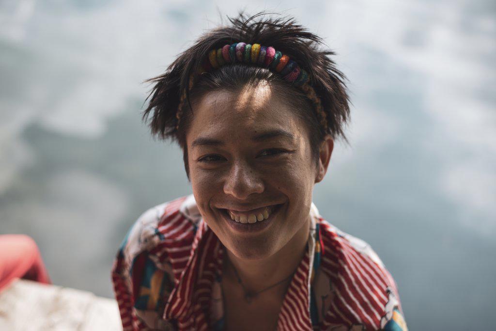 non-binary asian person smiles in colourful shirt by lake