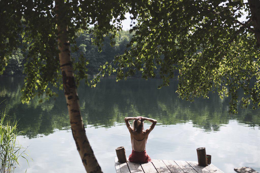 Young person relaxing lakeside on wooden jetty under trees in summer