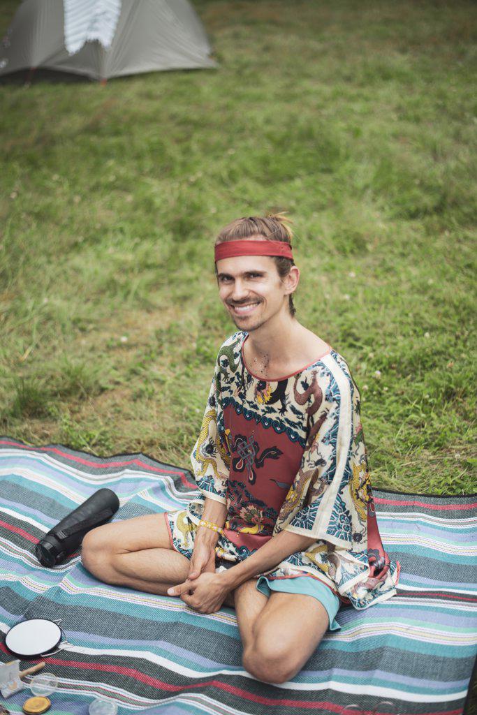 Smiling man in silk dragon top sits on picnic rug at camp on grass