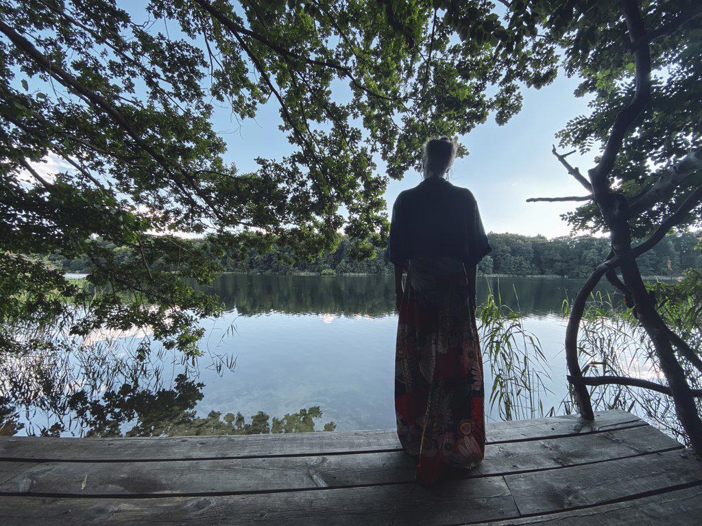 woman in skirt looks out from wooden platform over green natural lake