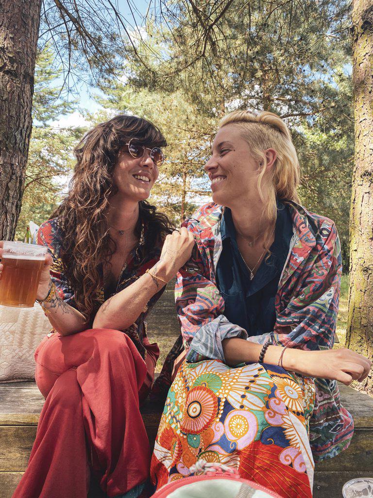 Happy queer women couple smile affectionately over beer in forrest