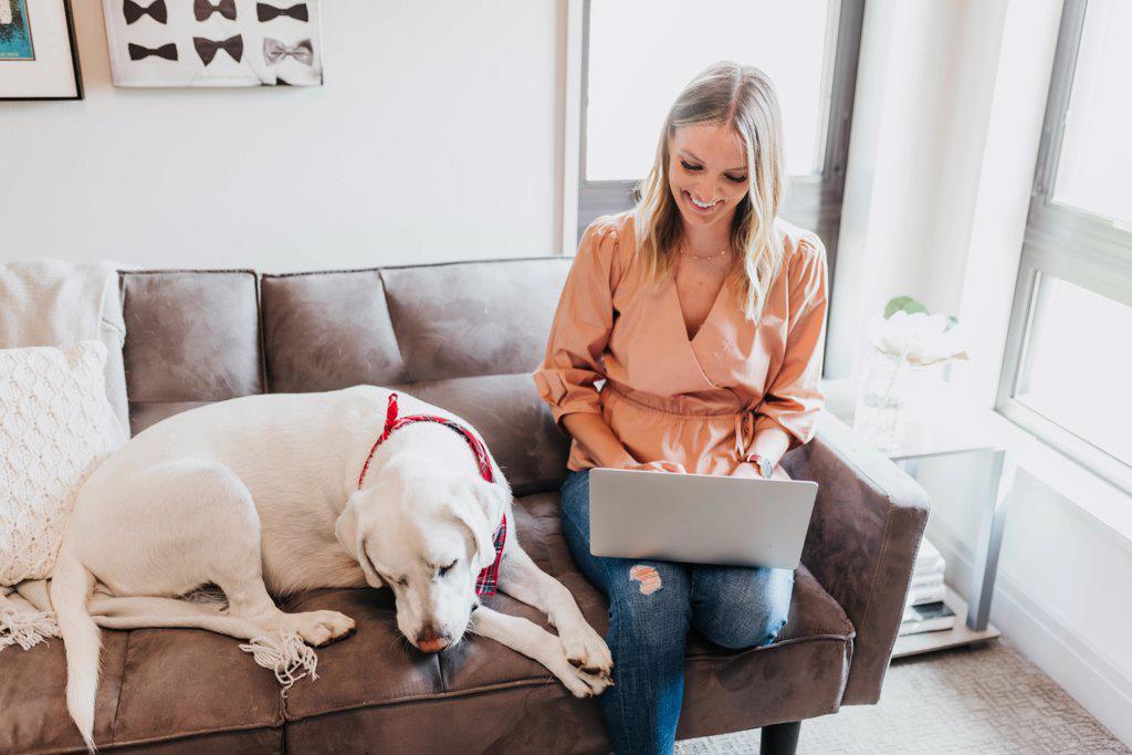 Woman works on laptop computer while sitting on couch with dog